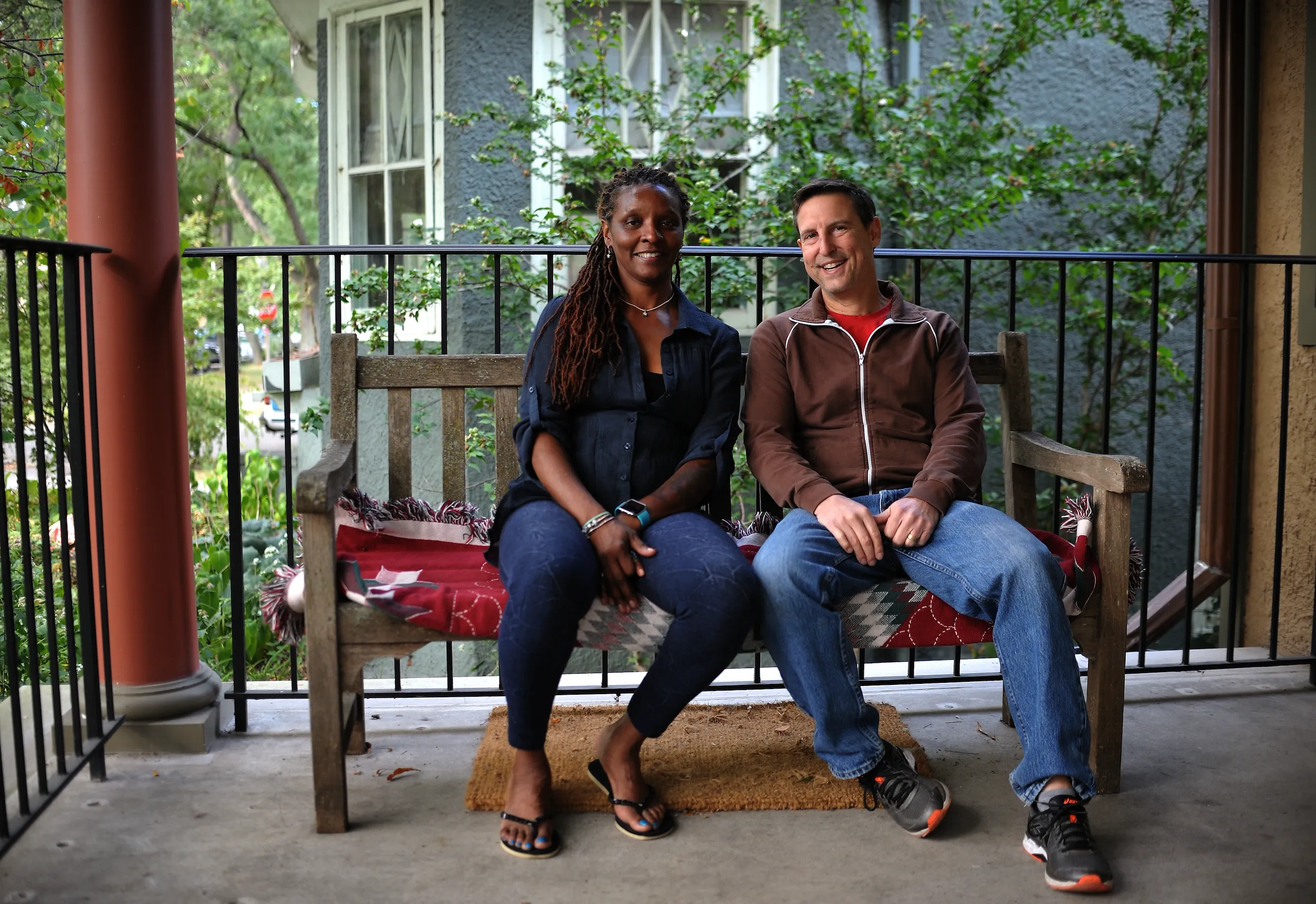 A Black woman and white man are sitting upon a wood bench on the porch of their home.